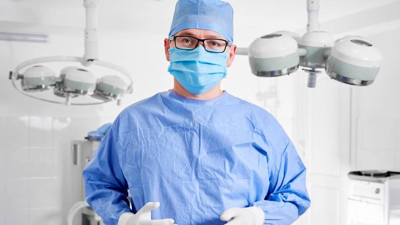 Gyno surgeon in sterile gloves, blue surgical uniform and protective face mask, ready for plastic surgery in clinic